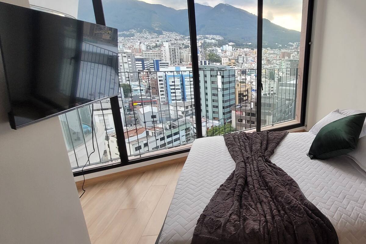 Quarto com uma cama grande, televisão fixada na parede e vista da cidade por uma janela ampla.