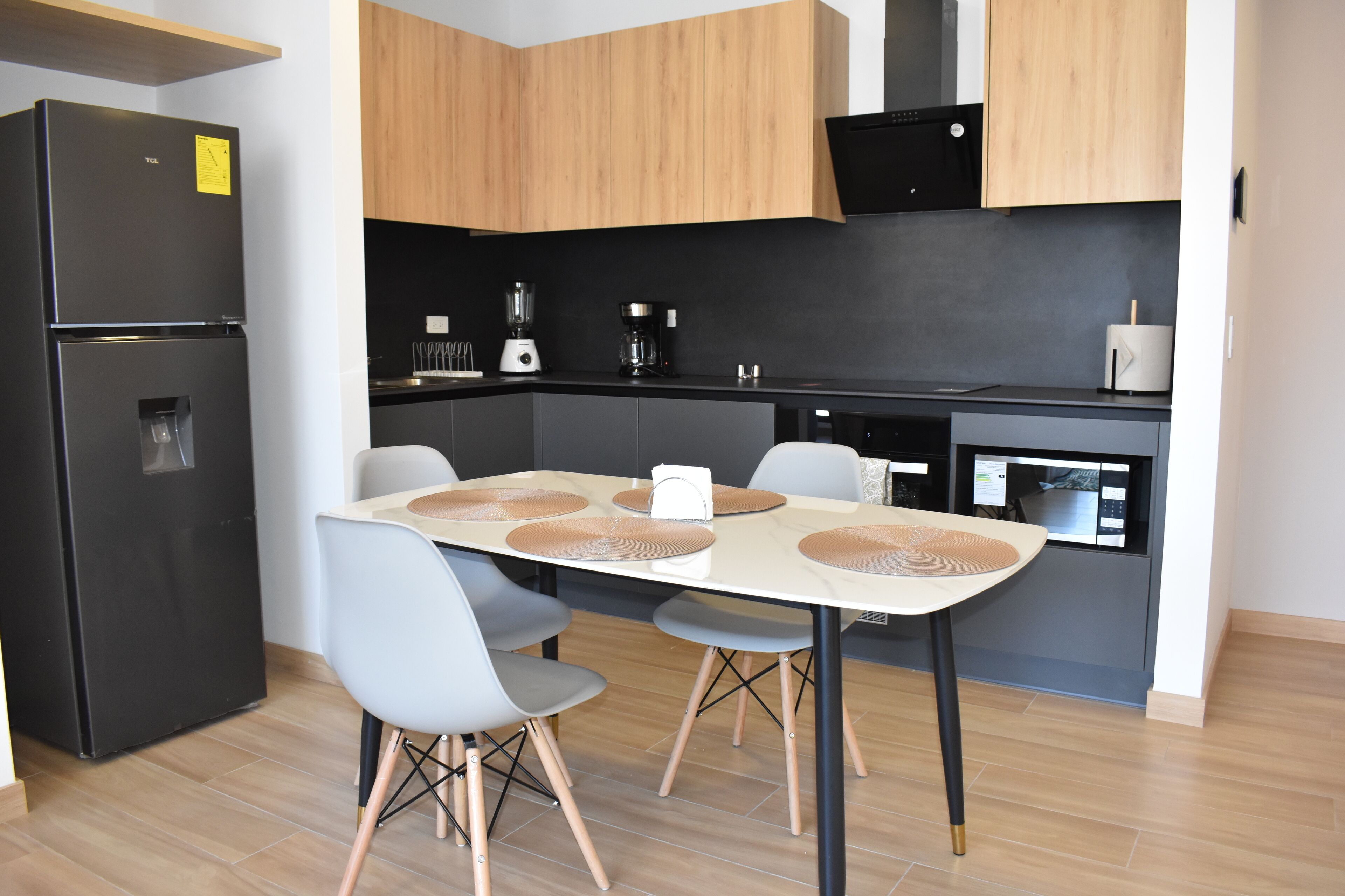 A modern kitchen with a black refrigerator, wooden cabinets, a dining table with chairs, and a microwave.