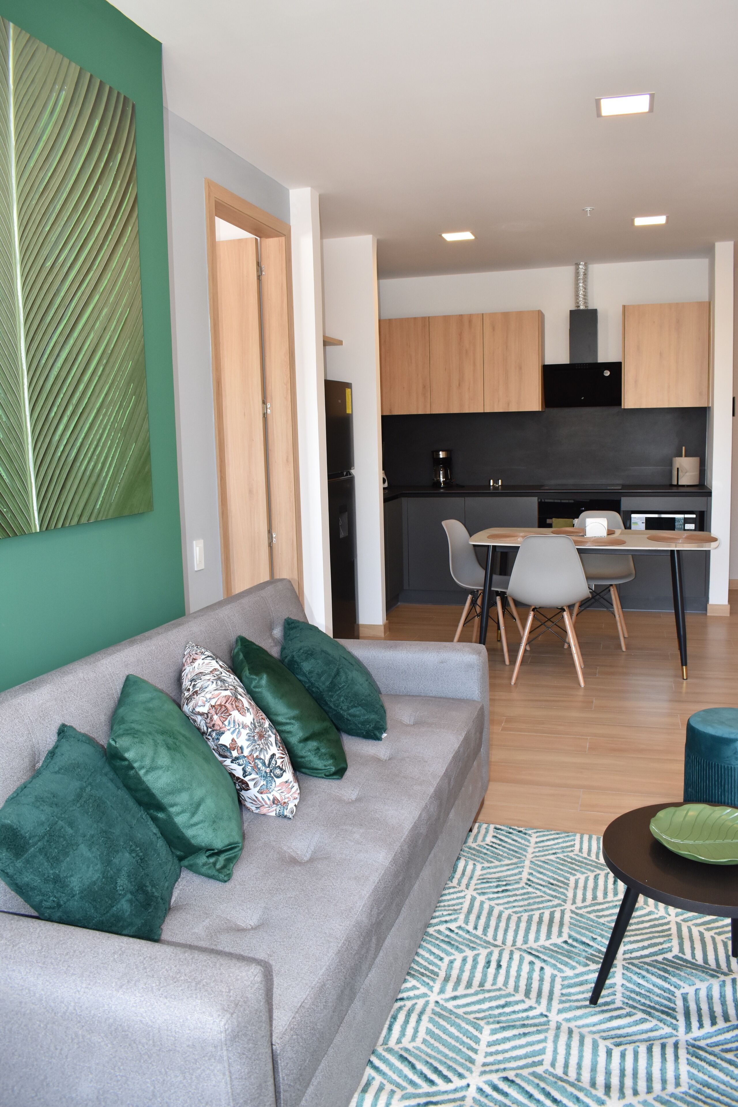 Un salon moderne avec un canapé gris, des coussins verts et un tapis à motifs. On aperçoit à l’arrière-plan un coin repas avec une table et des chaises.