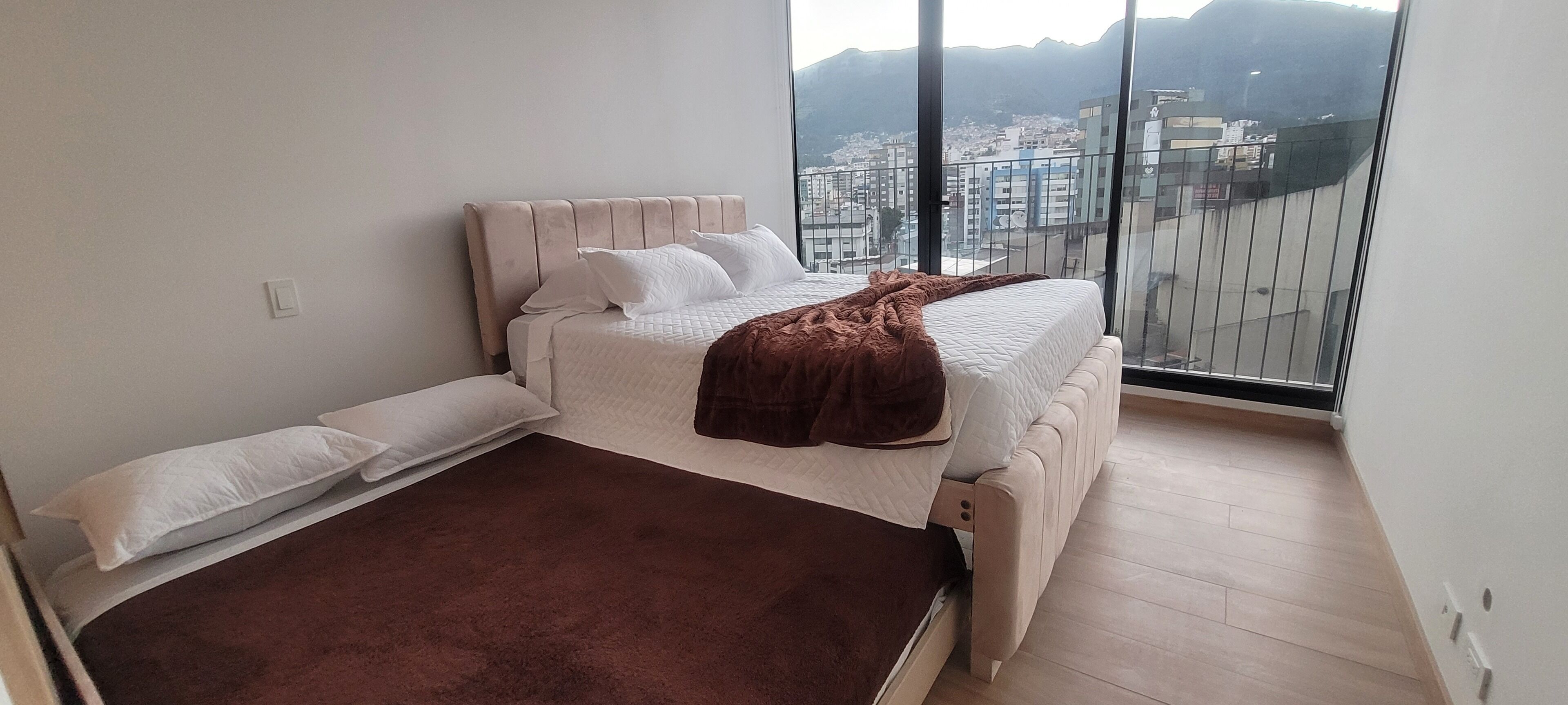 A bedroom with a bed, a brown blanket, and a city view through large windows.