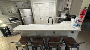 A kitchen with a large island, bar stools, a refrigerator, a microwave, and a fire extinguisher. - The Tiled Waters Retreat (Brandon)