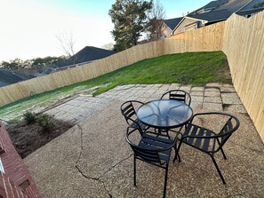 A patio area with a round table and four chairs, surrounded by a wooden fence and a grassy lawn.