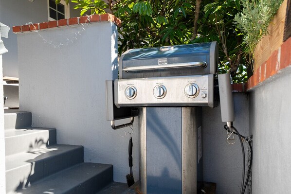 A Kenmore portable grill with three burners, mounted on a metal stand, against a wall with a brick baseboard and a window.