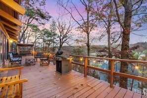 Una terraza de madera con mobiliario exterior, una parrilla y vistas a árboles y edificios.