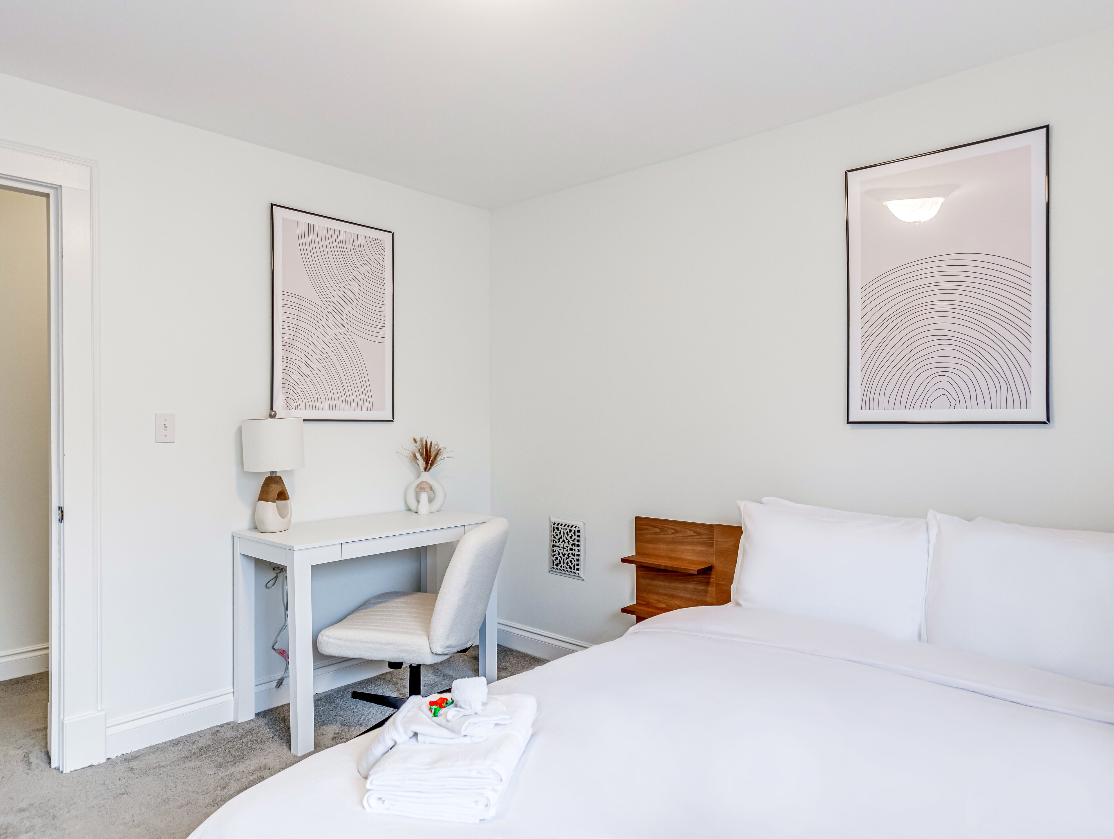 Ett hotellrum med en säng, ett skrivbord med en stol, två inramade abstrakta konstverk, ett nattduksbord med en lampa och en liten dekorativ vas.