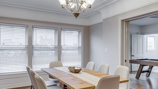 A dining area with a wooden table, six chairs, a chandelier, and large windows with blinds.