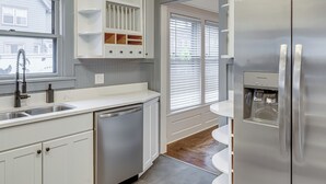 A modern kitchen with white cabinetry, a stainless steel dishwasher, a refrigerator, and a window with blinds.