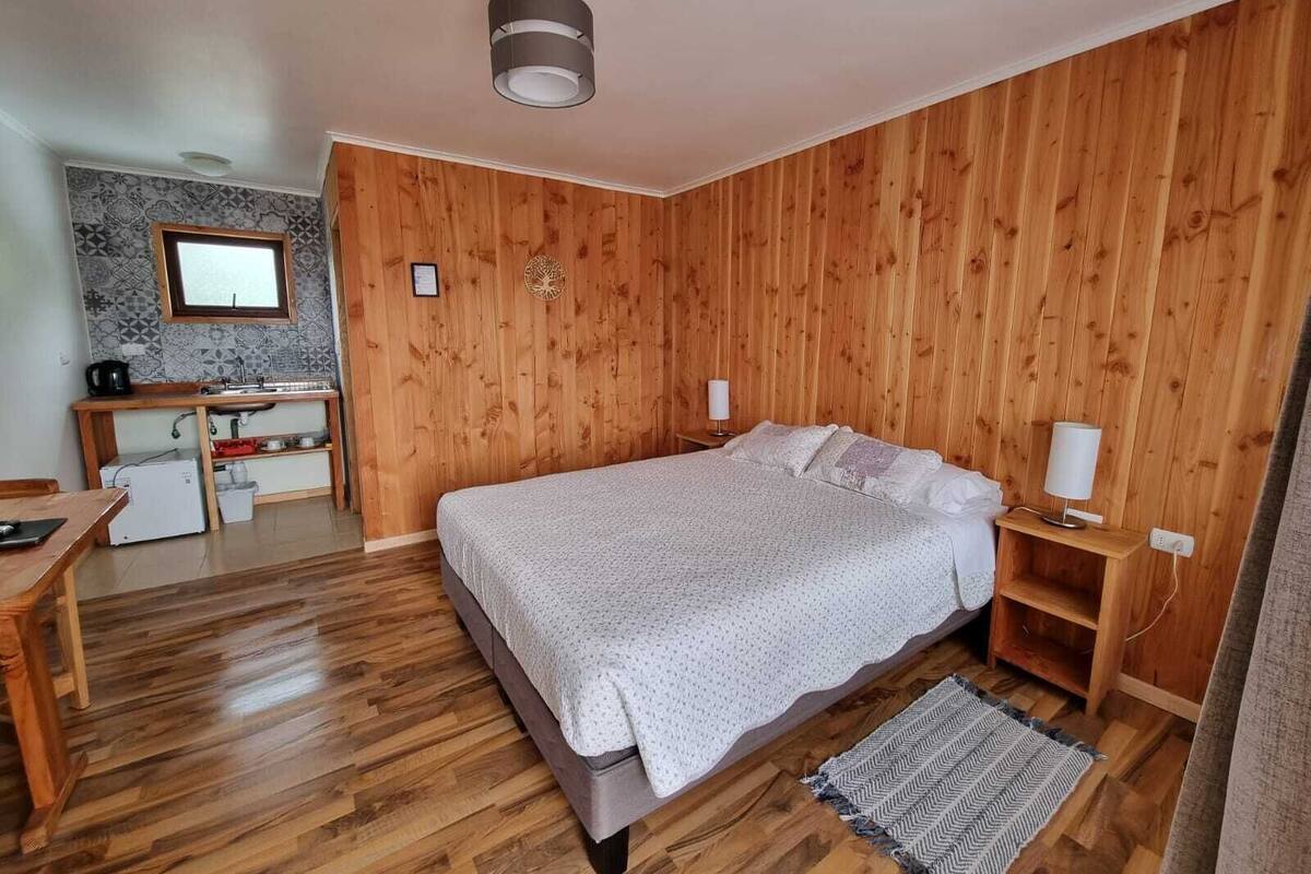 Een slaapkamer met een houten vloer, een bed met wit beddengoed, een houten bureau met een computer en een klein raam.
