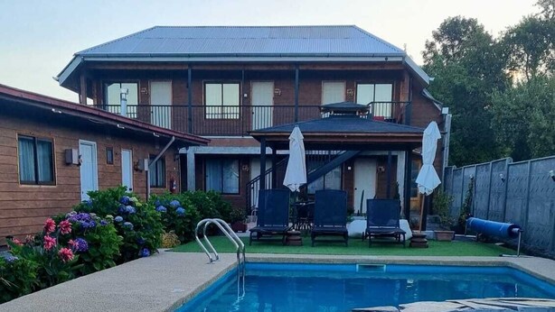 A swimming pool with a stone edge, a wooden house with a balcony in the background, and a grassy area with lounge chairs. - Antu Apart Hotel Villarrica (Villarrica)