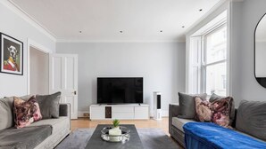 Un salón moderno con dos sofás grises, una mesa de centro con un adorno, un televisor de pantalla plana sobre un mueble blanco y un ventanal que deja entrar luz natural.