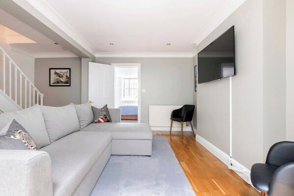 A modern living room with a sectional sofa, a flat-screen TV mounted on the wall, and a wooden floor.