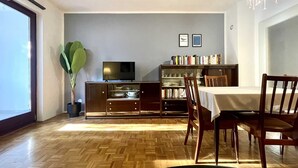 A dining area with a wooden herringbone floor, a dining table with chairs, a television on a cabinet, and a bookshelf filled with books.