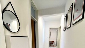 A hallway with grey tiled flooring, white walls, and a door leading to another room. There are framed pictures on the wall and a mirror on the left.