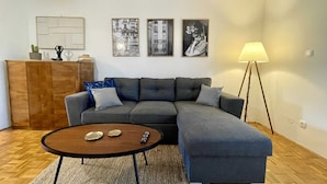 A modern living room with a grey sectional sofa, a wooden coffee table, and a floor lamp. There are three framed black and white photographs on the wall.