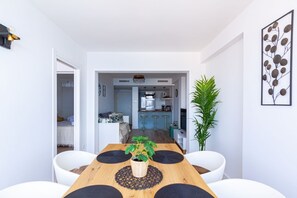 A modern dining area with a wooden table, black placemats, and a potted plant. A kitchen is visible in the background with a bar counter and stools.