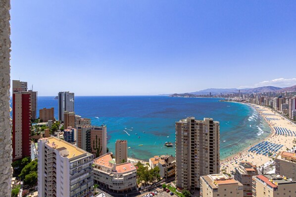 A coastal cityscape with high-rise buildings, a beach with umbrellas, and clear blue waters.