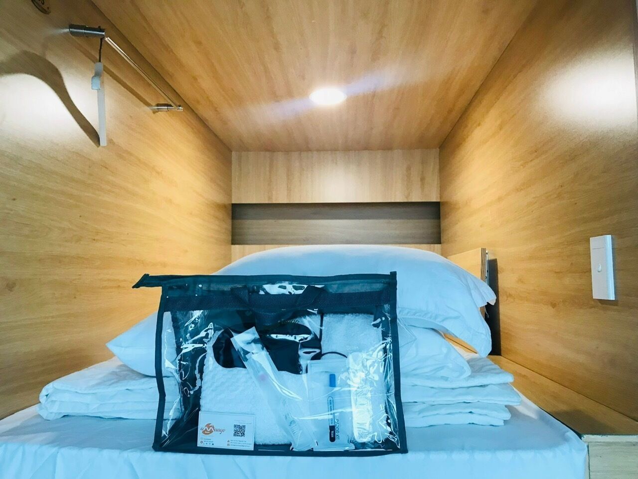 A small, narrow hotel room with wooden walls and a single bed. There is a bag of toiletries on the bed.