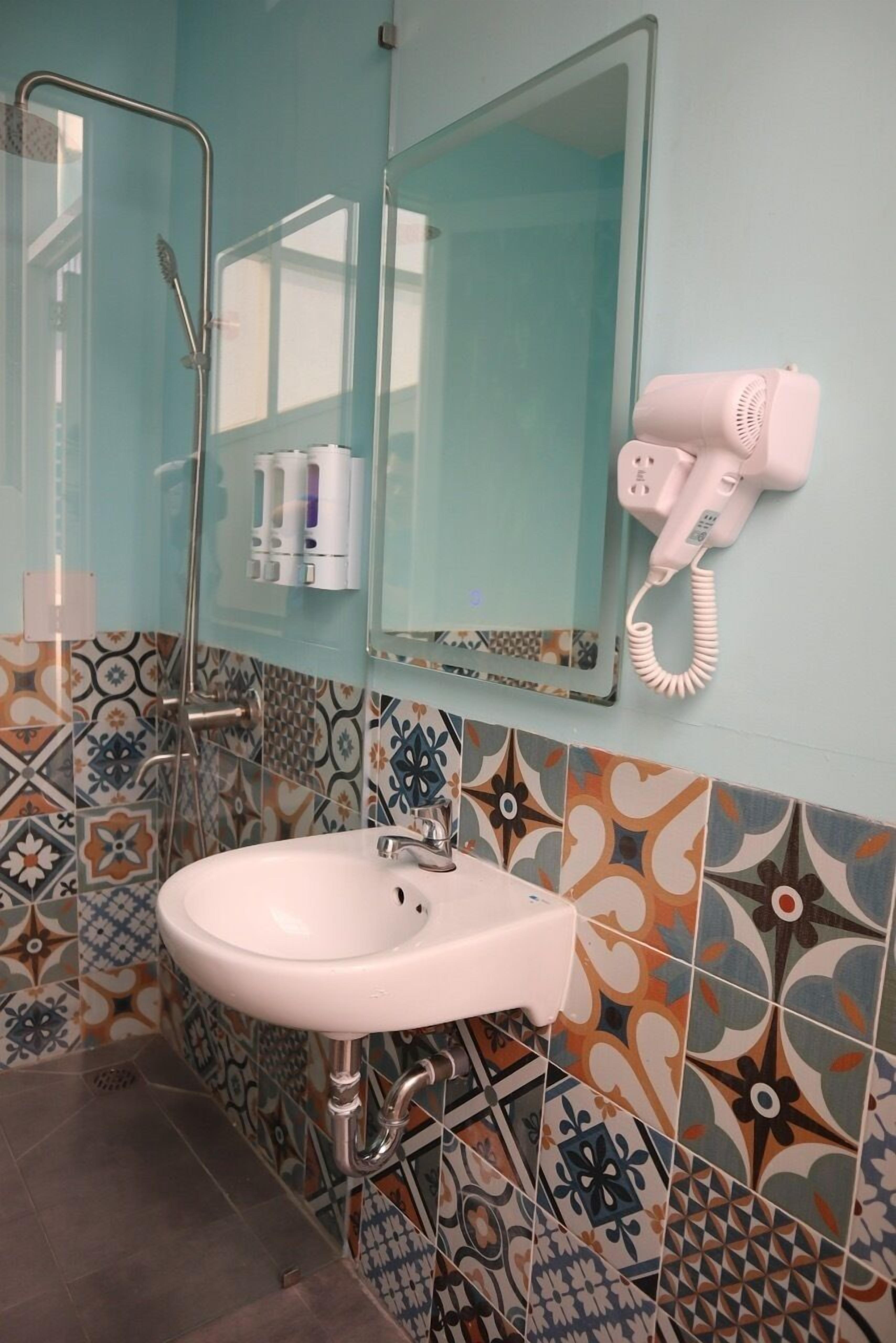 Un baño con un lavabo blanco, cabina de ducha de vidrio, secador de pelo rosa y azulejos estampados.