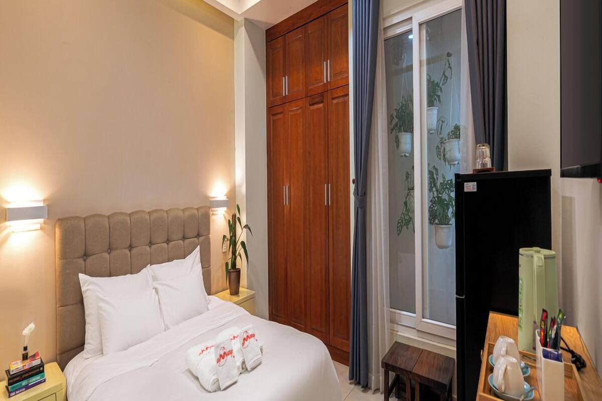 A hotel room with a bed, a ceiling fan, a wardrobe, a desk with a chair, and a window with curtains.