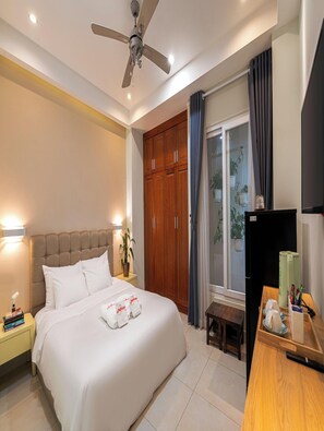 A hotel room with a bed, a ceiling fan, a wardrobe, a desk with a chair, and a window with curtains.