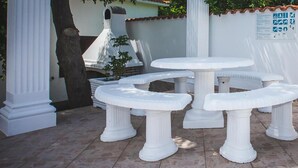 Un espacio de patio con mesas y columnas de piedra blanca, un muro de ladrillo y suelo de baldosas.