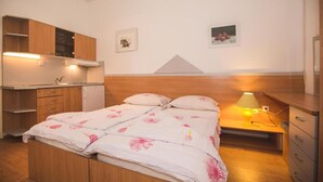 Un dormitorio con cama, una mesita de noche con lámpara, un escritorio pequeño, kitchenette y un estante en la pared.