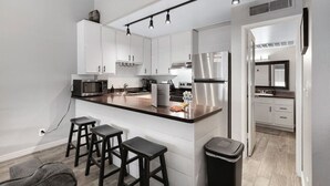 Cozinha moderna com armários brancos, geladeira de aço inoxidável, micro-ondas e cafeteira. Há três banquetas e uma lixeira.