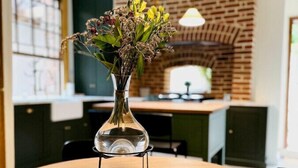 Una mesa redonda de madera con un florero de vidrio transparente que contiene flores secas, una pared de ladrillo y una lámpara colgante en una cocina moderna.