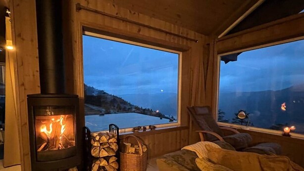 Interior acogedor de una cabaña con estufa de leña, un sofá, una silla y un amplio ventanal con vistas a un lago al atardecer.