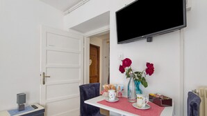 A small room with a white door, a flat-screen TV mounted on the wall, a table with a red placemat, two chairs, and a vase with red flowers. - Room 'Abeti' with Mountain View (San Pietro di Cadore)