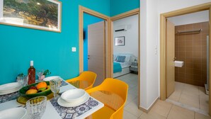 Un coin repas avec une table dressée pour deux personnes, un tableau encadré au mur et une vue sur une chambre à coucher avec un lit et une table de chevet.