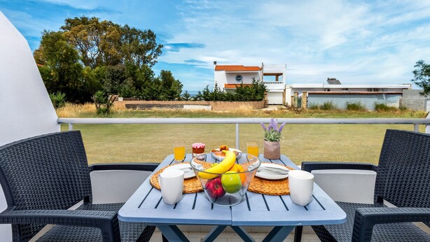 Uma área de refeições ao ar livre com uma mesa posta para dois, incluindo uma cesta de frutas, pratos, talheres e um copo de suco de laranja.