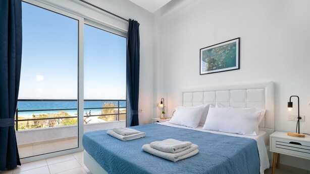 A bedroom with a large bed, a nightstand, a bedside table, a framed picture on the wall, and a view of the ocean through a sliding door. - Apartment 'Rhodes Sea Ialysos' with Balcony, Wi-Fi and Air Conditioning (Ialisos)