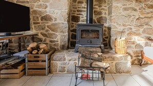 Una habitación con paredes de piedra, un estufa de leña, un televisor y una mesa con libros y una computadora portátil.