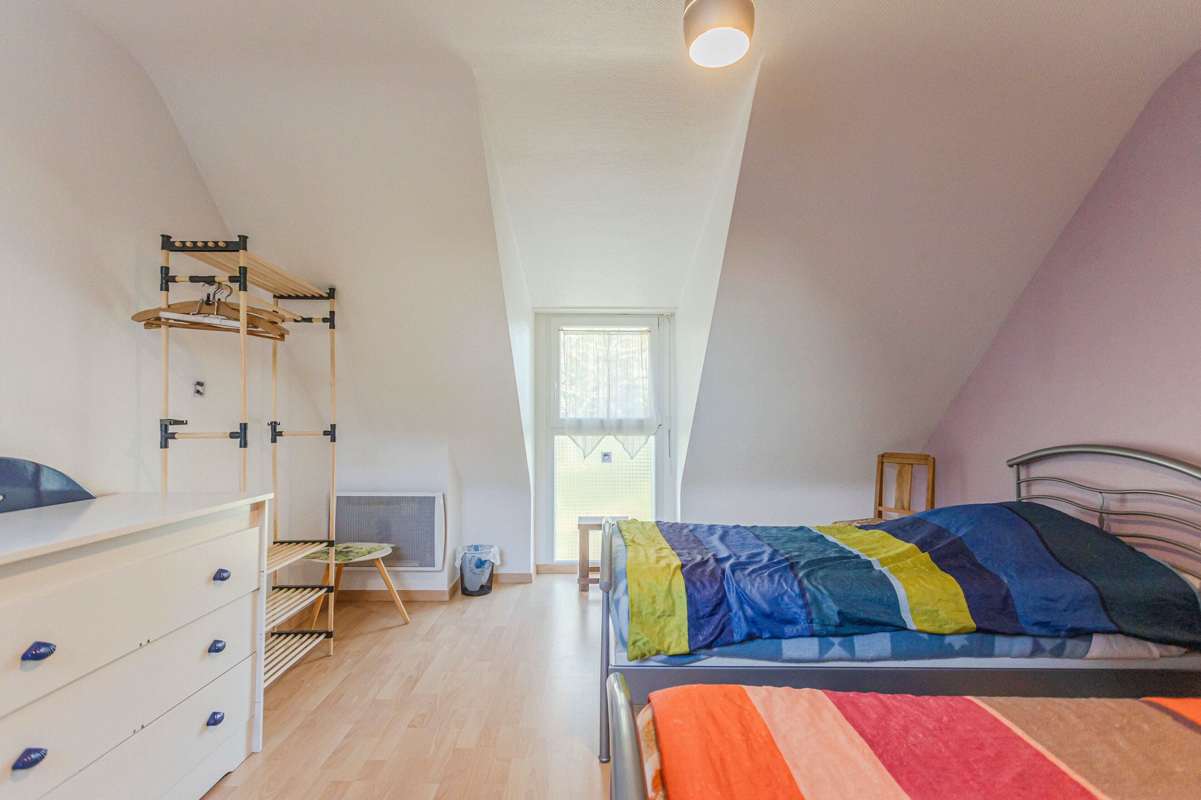 Un dormitorio con cama, cómoda, radiador, ventana y perchero.