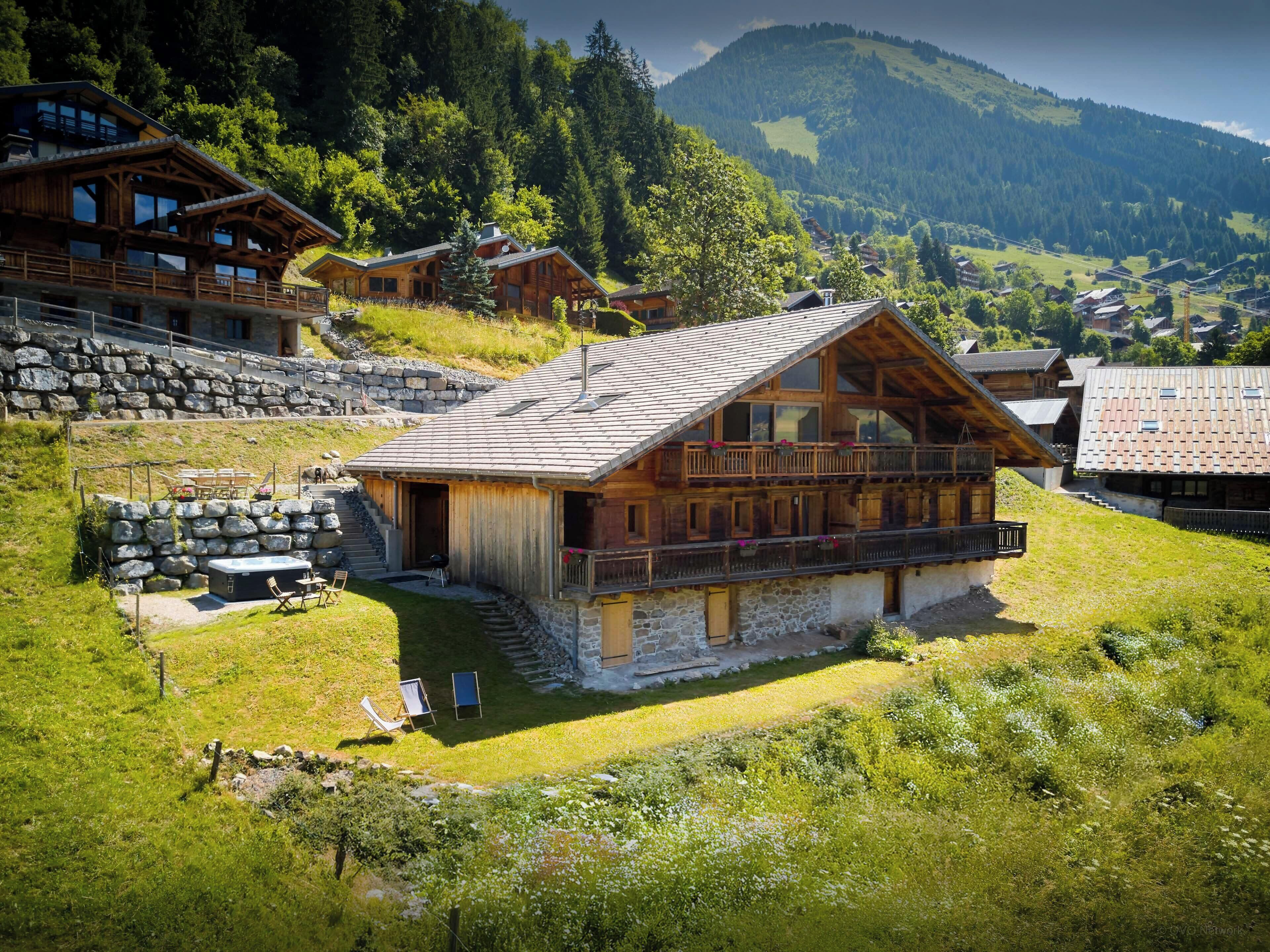 A wooden chalet with a stone foundation, a balcony, and a view of a mountainous landscape.
