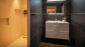 Un baño moderno con paredes de baldosas oscuras, un tocador blanco con espejo e iluminación empotrada en el techo.