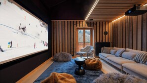 A cozy living room with a large screen displaying a ski event, a sofa, two poufs, and a wooden-paneled wall.