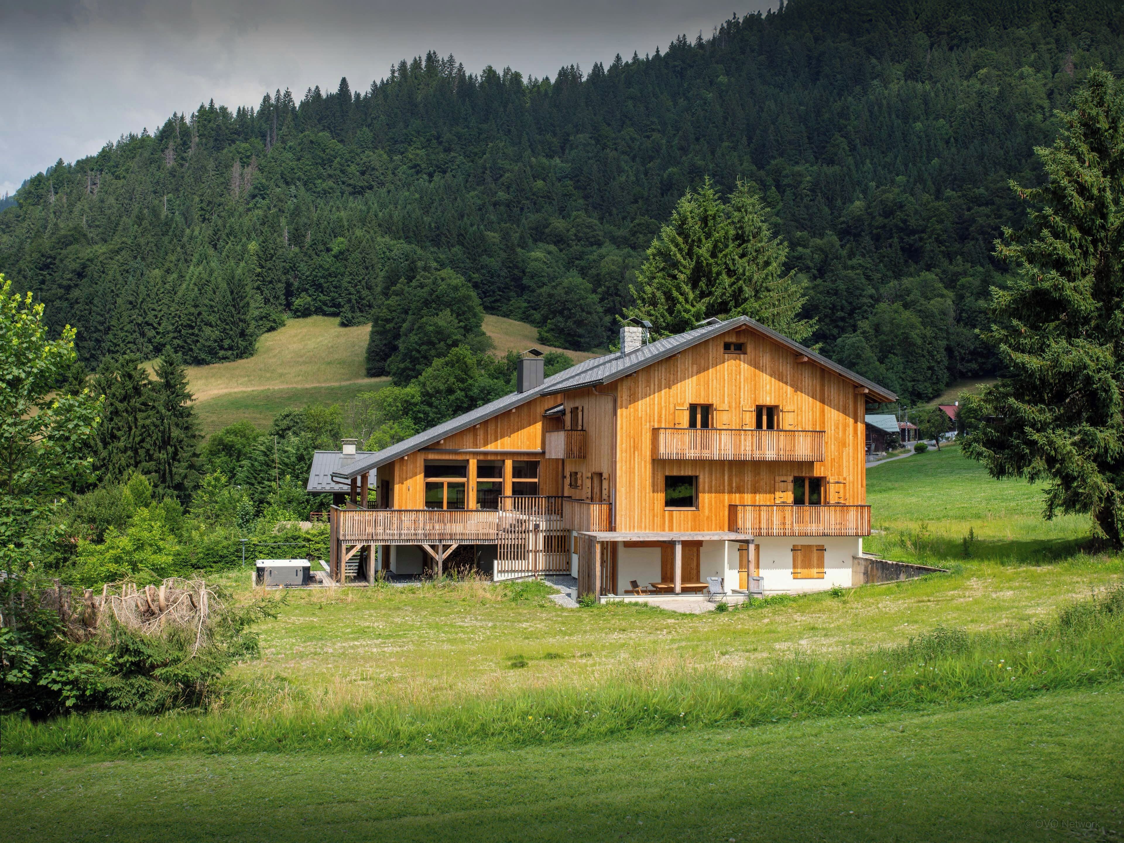 A wooden cabin with a balcony, surrounded by a grassy field and dense forest.