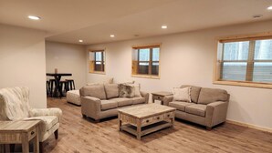 A spacious living room with a wooden floor, two sofas, a coffee table, and a chair. There are windows with blinds, a dining table with chairs, and a small table with a lamp. - Spacious 5-BR home in Manitowish Waters with pier access on Chain of Lakes. (Manitowish Waters)