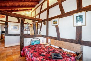 A room with exposed wooden beams, a bed with a floral bedspread, a wooden bench, and a small table with a lamp.