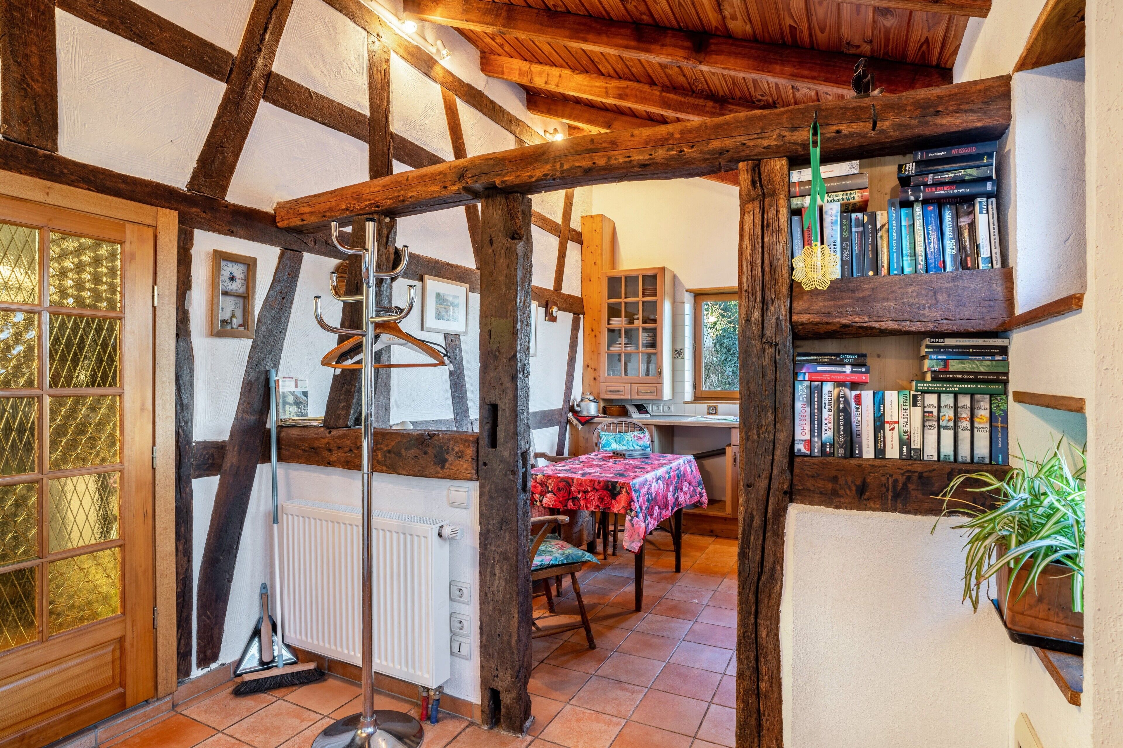 A rustic interior with exposed wooden beams, a dining area with a table and chairs, a radiator, a bookshelf, and a hanging plant.