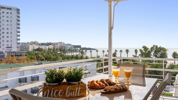 A balcony with a table set for breakfast, including croissants and orange juice, overlooking a cityscape with palm trees and buildings. - Apartment 'Mas' with Sea View, Wi-Fi and Air Conditioning (Salou)