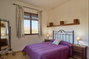A bedroom with a bed, two bedside tables with lamps, a mirror, a window with curtains, and a shelf with baskets. - Country House 'Casa Rural El Rincon' with Mountain View, Wi-Fi and Air Conditioning (Carcabuey)
