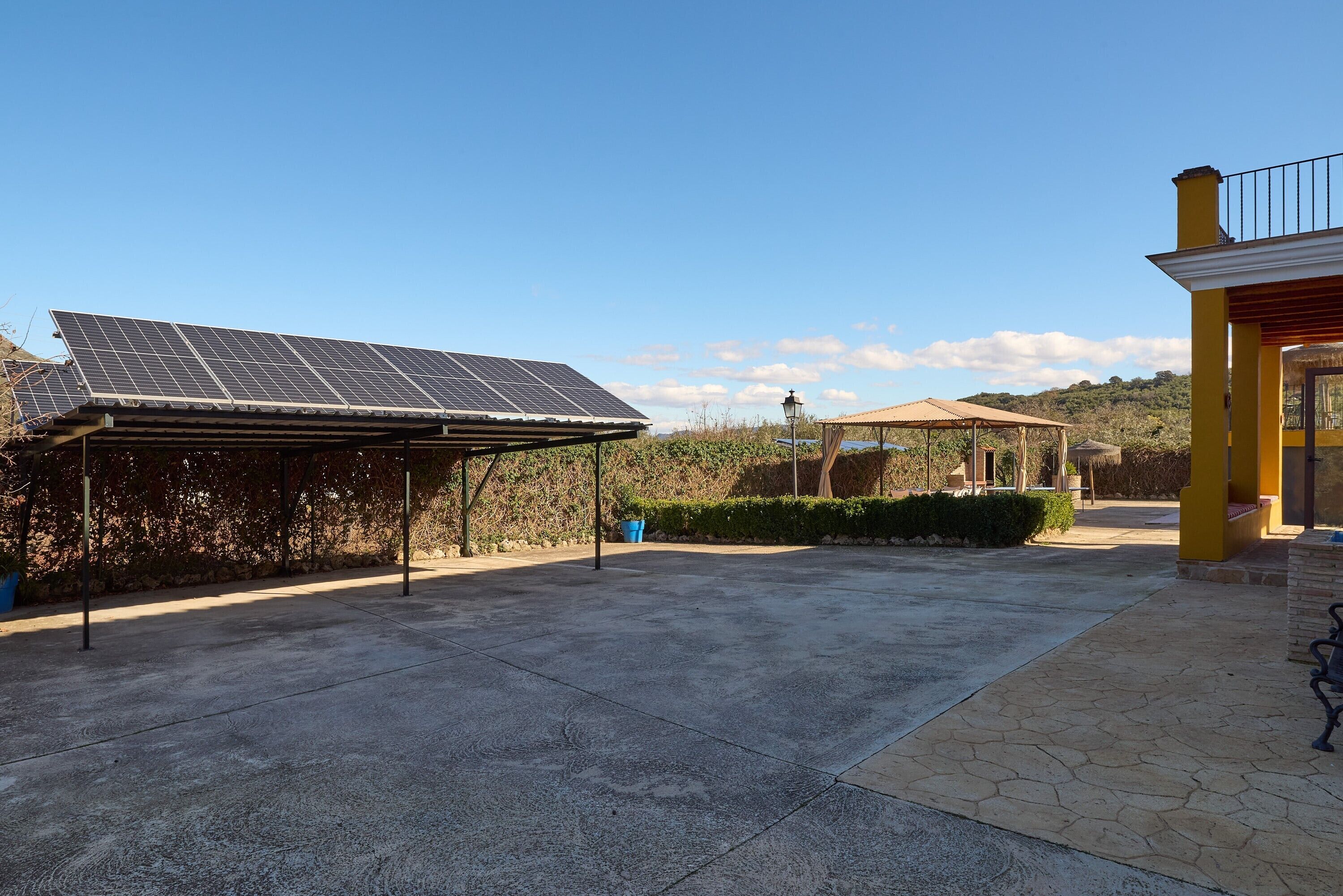 A covered outdoor area with solar panels, a concrete patio, and a building with a yellow facade.