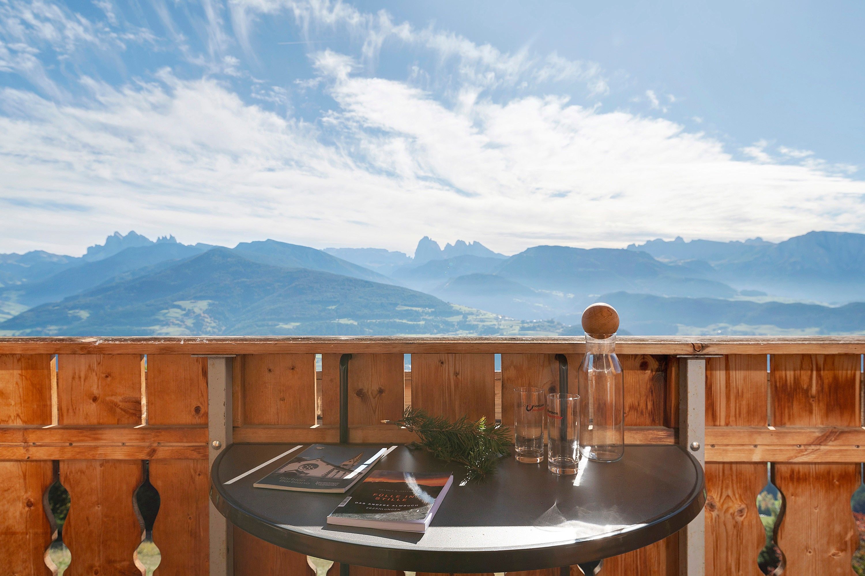 Una terraza de madera con una mesa que sostiene libros y gafas, con vistas a un paisaje montañoso.
