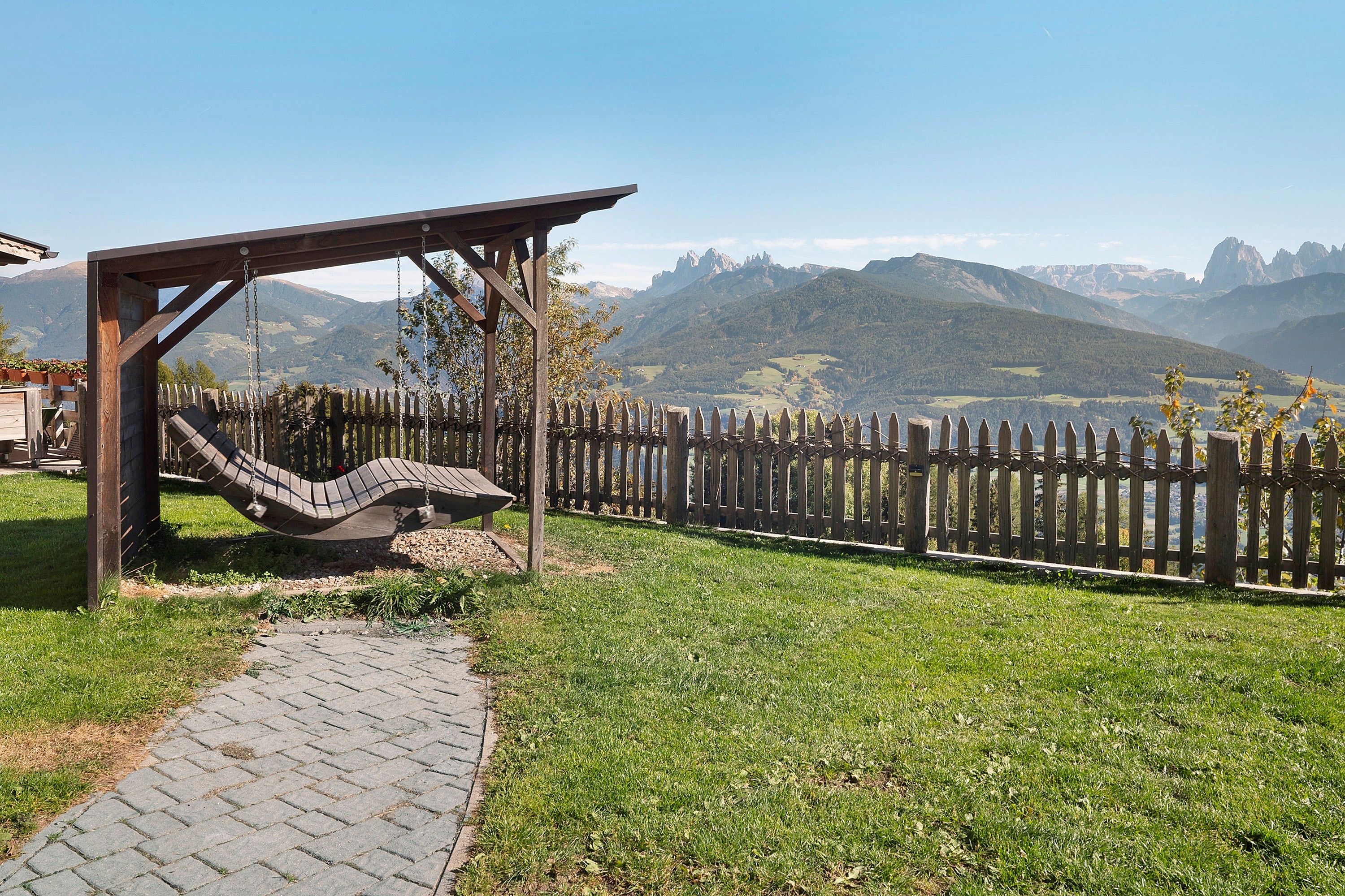 Una pérgola de madera con una hamaca, un sendero pavimentado y una cerca de madera con vistas a un paisaje montañoso.