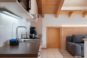 A compact kitchen with a sink, stove, and coffee maker, a wooden door, and a grey sofa.