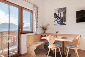 A room with a wooden bench, a table, and chairs, a window with curtains, and a black and white photograph on the wall.
