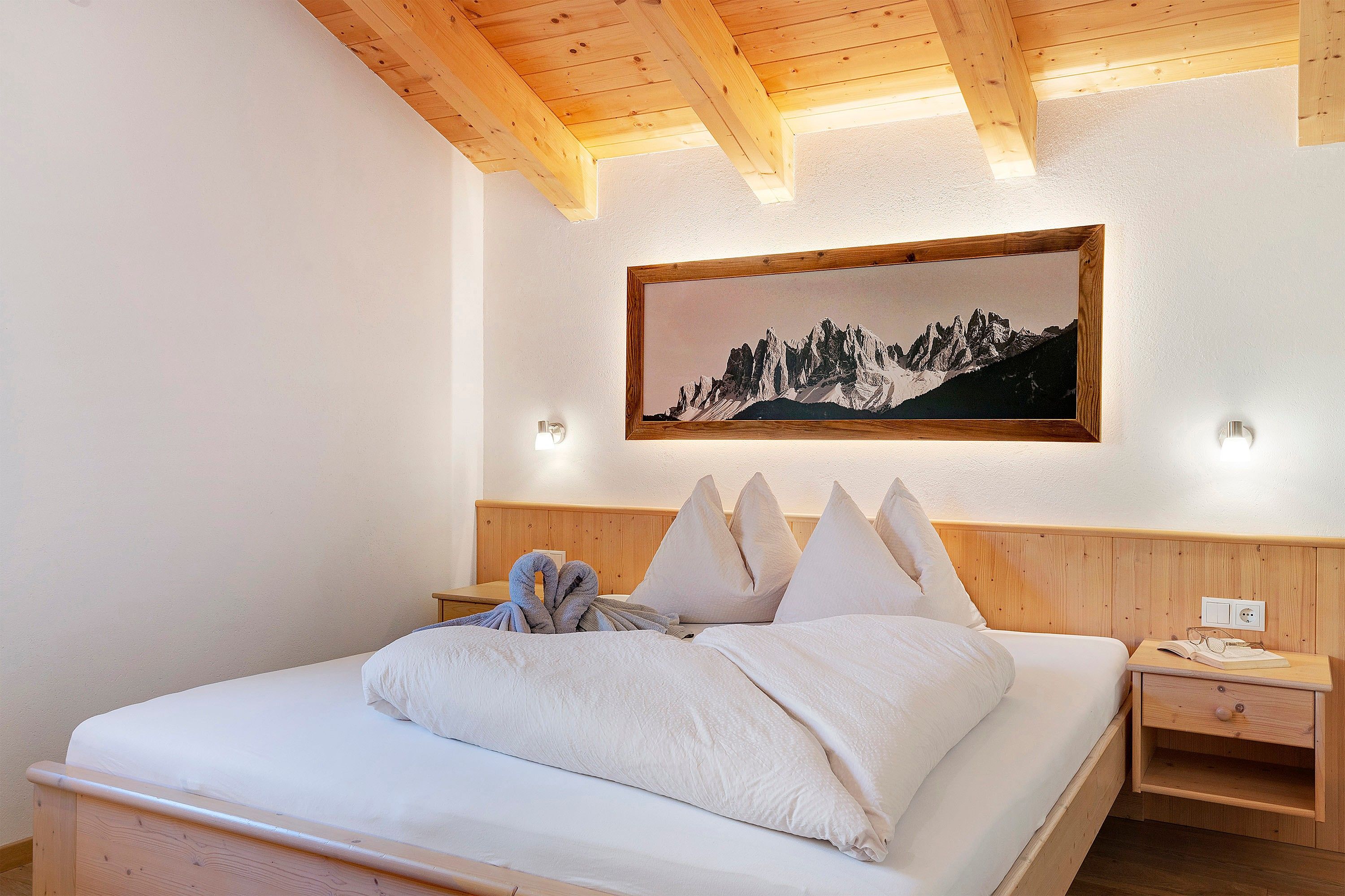 Un dormitorio con cabecera de madera, cama con ropa de cama blanca, un cuadro de un paisaje montañoso y una mesita de noche con una lámpara.